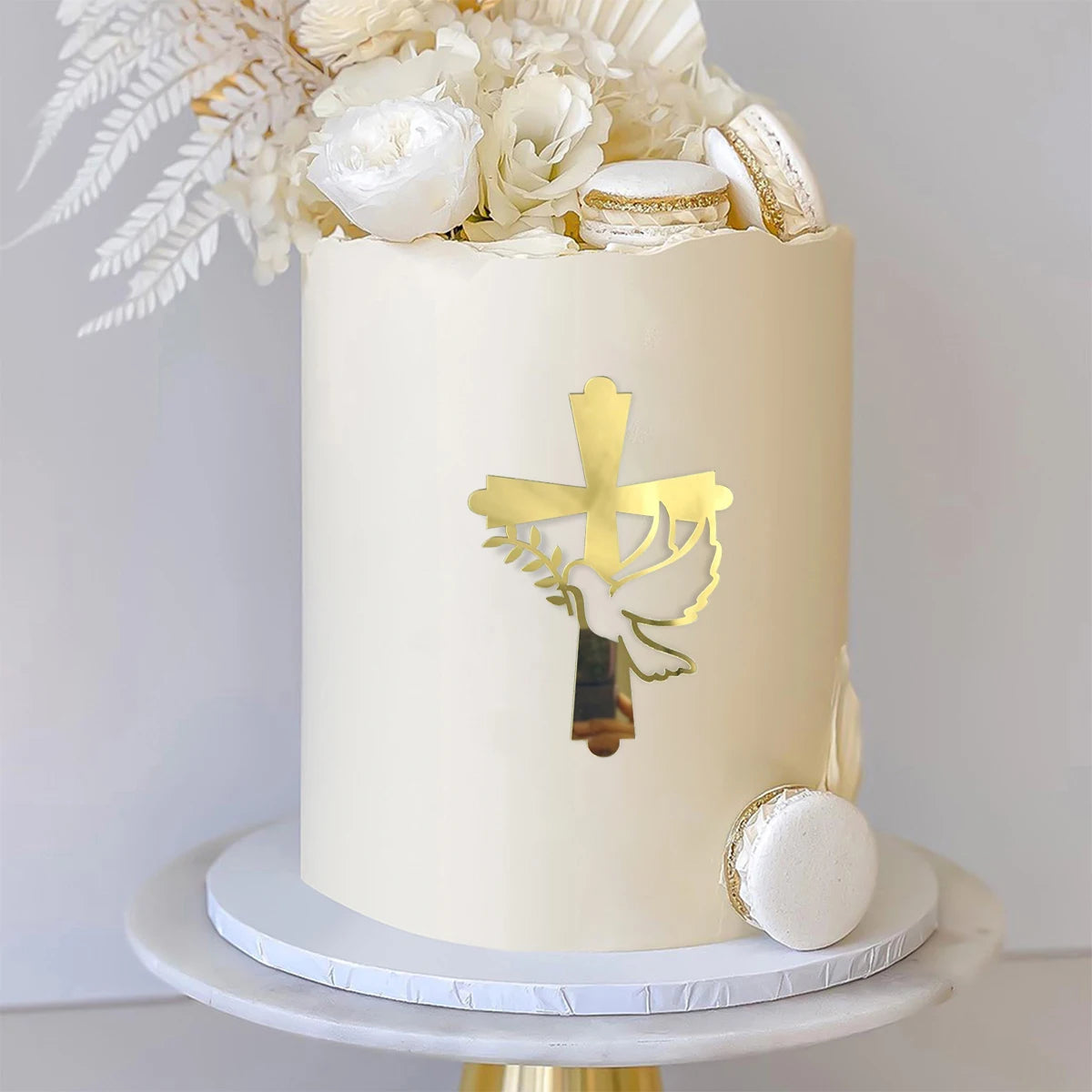 A round cream-colored cake decorated with a gold acrylic cross topper and white floral accents.