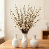 A vase holds a spray of white artificial plum blossoms on a wooden table.
