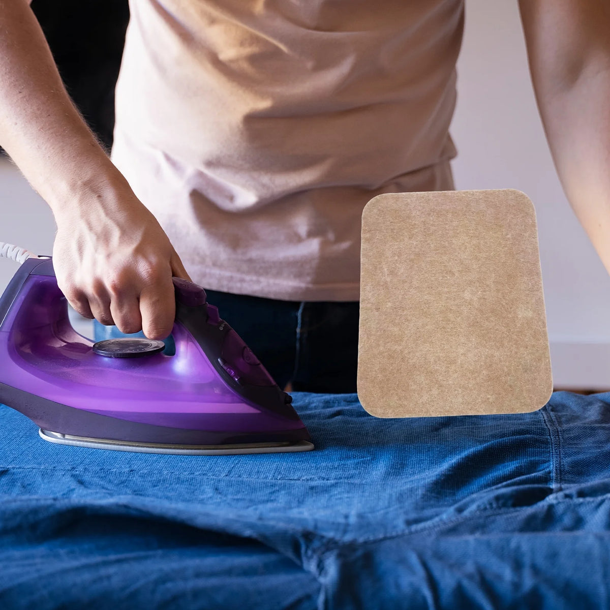 A hand is ironing fabric on a blue shirt using a purple iron and a brown felt ironing mat.
