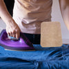 A hand is ironing fabric on a blue shirt using a purple iron and a brown felt ironing mat.