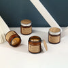 A brown glass jar candle with a silver lid sits on a white surface next to a candle in a similar jar.