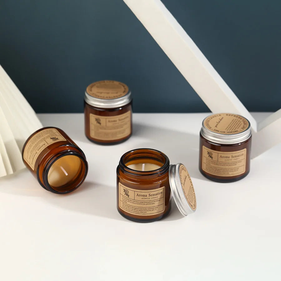 A brown glass jar candle with a silver lid sits on a white surface next to a candle in a similar jar.