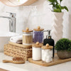 A collection of white and purple bath products stored in glass jars with wooden lids on a light countertop.
