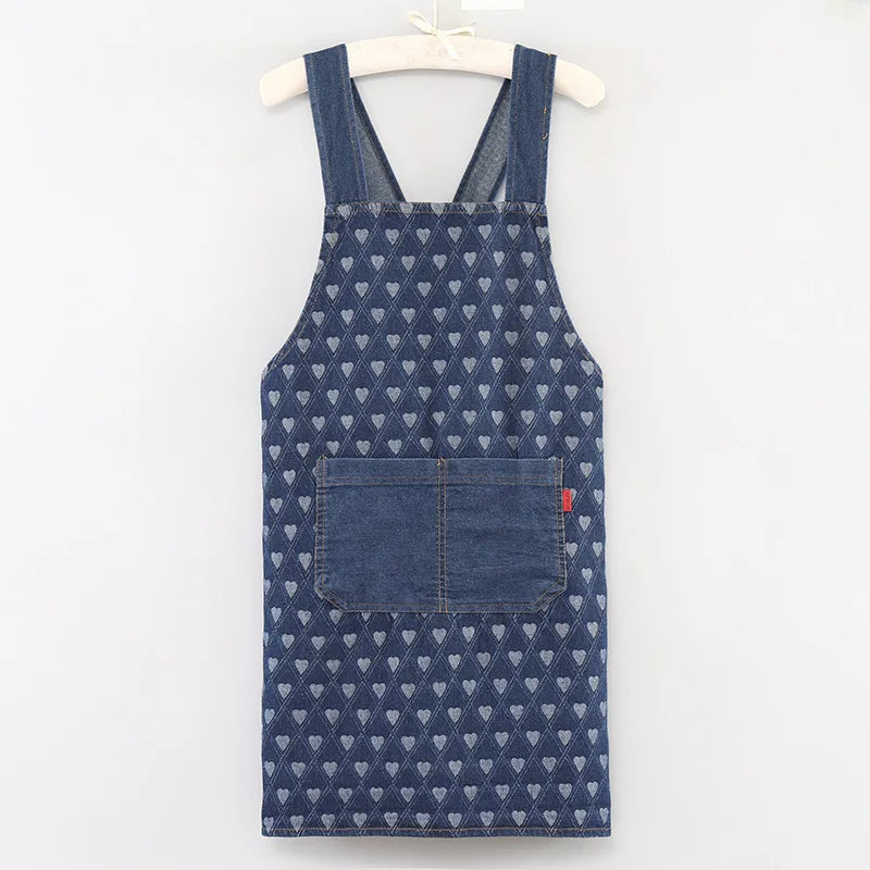 A dark blue denim apron with a repeating pattern of small white hearts and a chest pocket hanging on a wooden hanger.