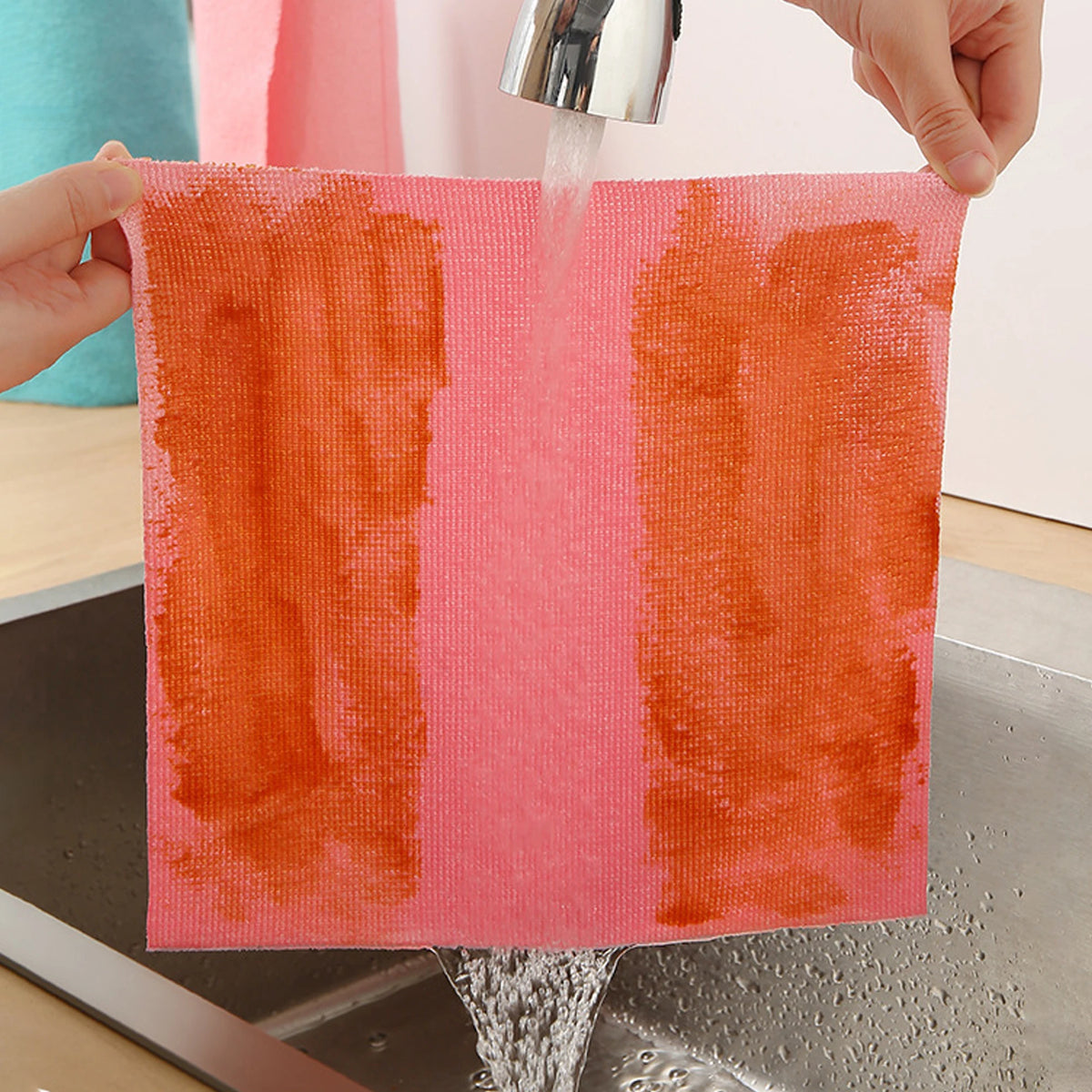 A hand is rinsing a bright coral-orange reusable microfiber cleaning cloth under running water.