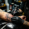 A tattoo artist wearing black nitrile gloves is applying ink to a client's arm.