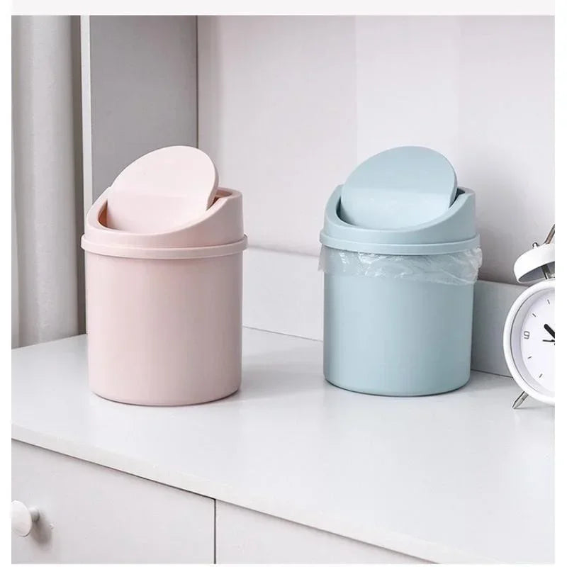 A pink compact desktop trash can with a light blue flip cover sitting on a white surface.