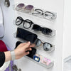 A hand places sunglasses and eyeglasses onto a wall mounted acrylic display shelf.