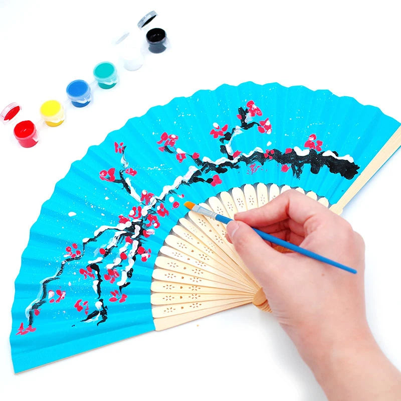 Une main peint un éventail en papier bleu vif avec des motifs floraux rouges et blancs.