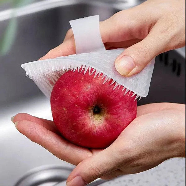 A hand holds a red apple under a white silicone kitchen cleaning brush for tough grime.