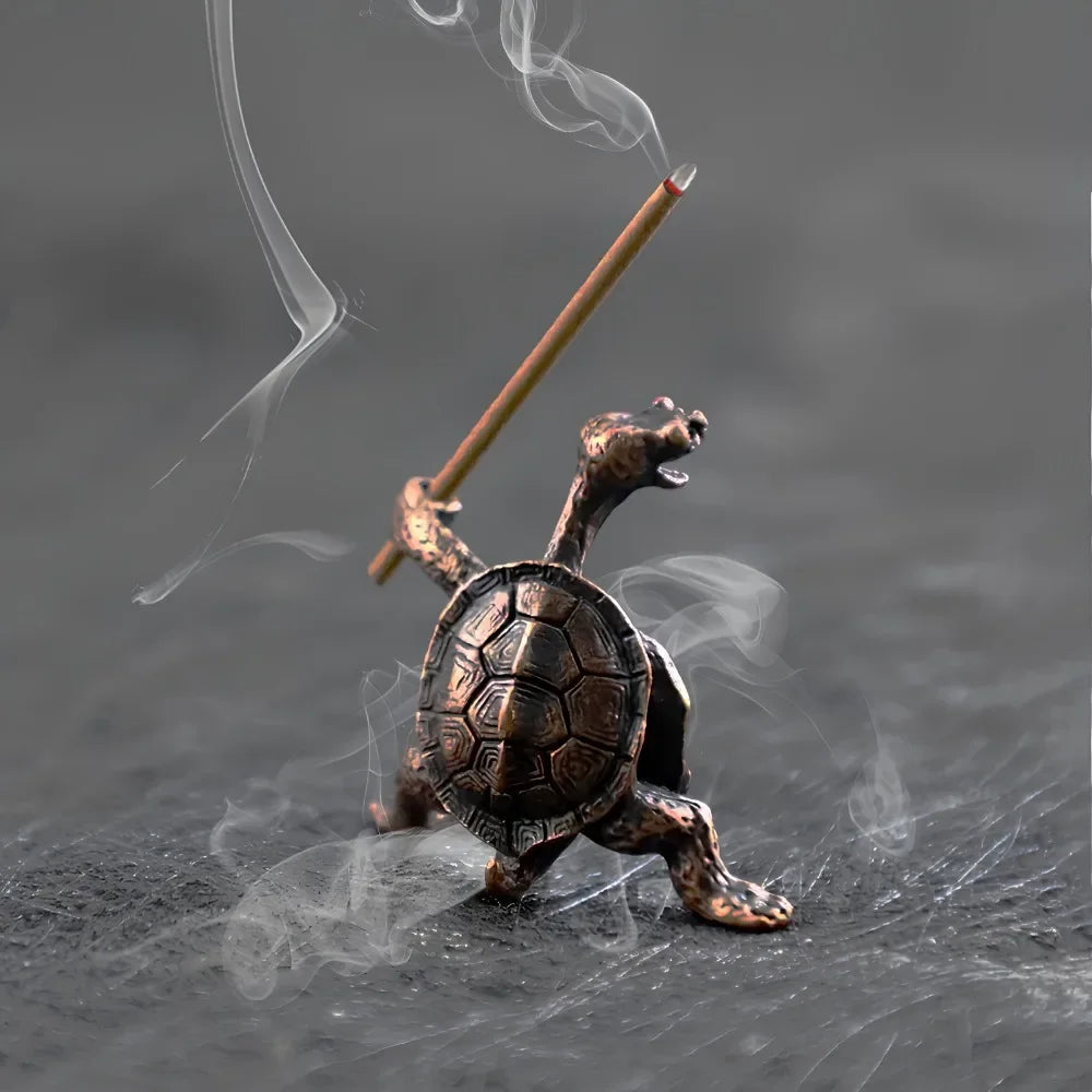 Une petite figurine de tortue en métal bronze avec de la fumée s'élevant d'un bâton, sur fond gris foncé.