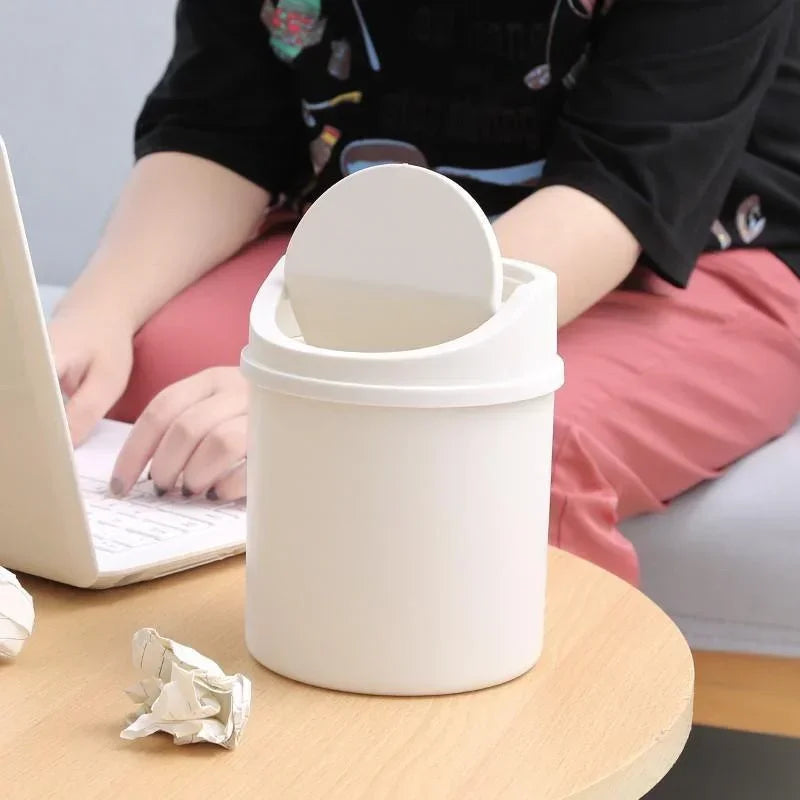 A white compact desktop trash can with a flip cover sits on a wooden table next to a laptop.