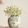 A hand holds a white ceramic bud vase filled with yellow and white wildflowers.