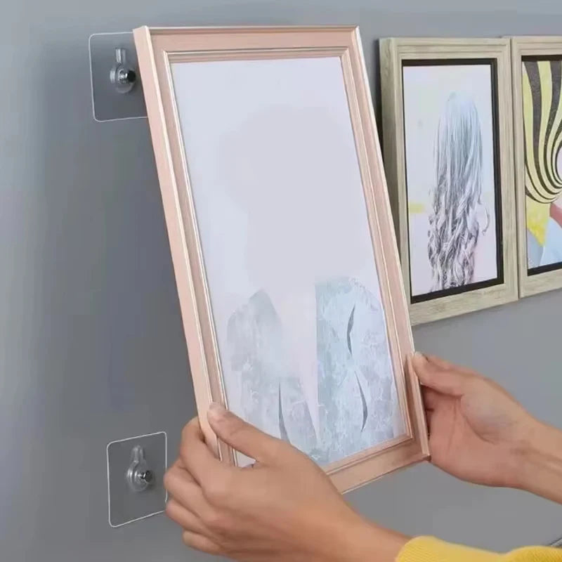 Hands are hanging a framed picture with a light pink frame onto a gray wall using adhesive hooks.
