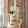 A tiered wedding cake decorated with white frosting, gold rhinestone toppers featuring a Mr & Mrs motif, and soft floral acce