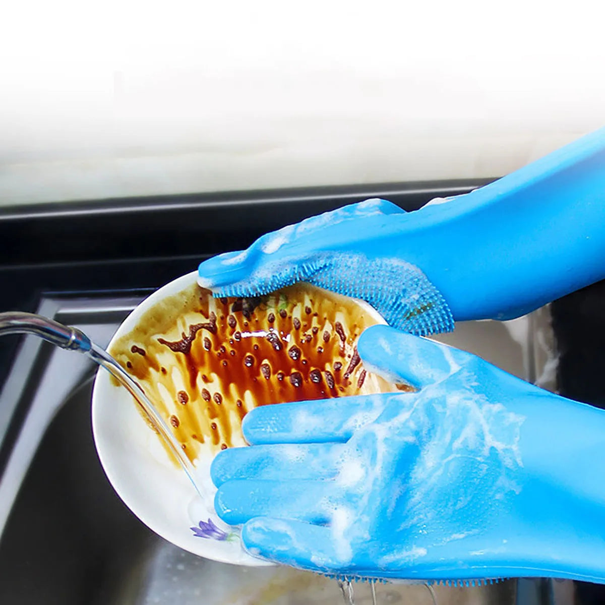 A hand wearing a blue silicone dishwashing glove is washing a plate with kitchen scrubber.