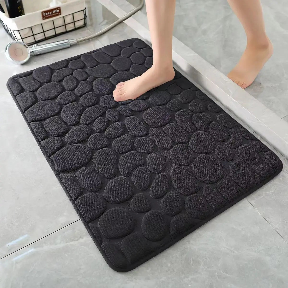 A black, textured 3D non-slip bathroom doormat placed on a light gray floor.