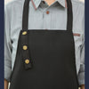 A black canvas apron with adjustable straps is worn over a light blue collared shirt.