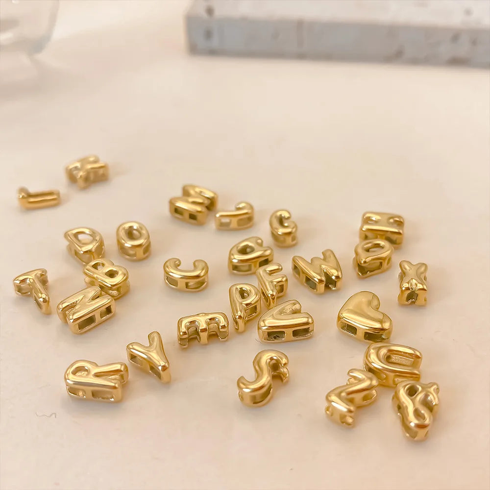 Eine Sammlung von goldvergoldeten 3D-Blasenbuchstaben-Anhängern, verstreut auf einer weißen Oberfläche.