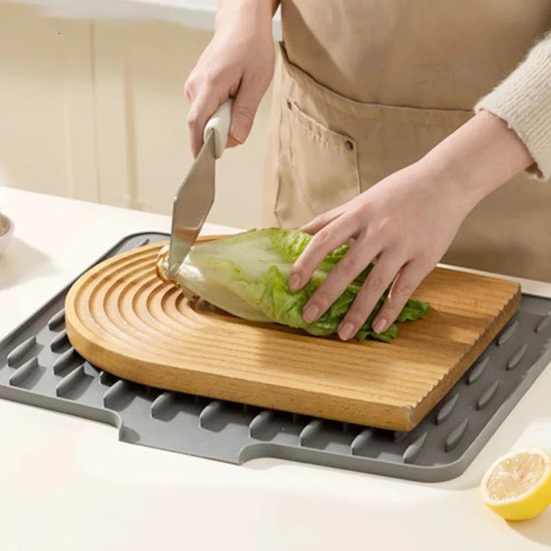 A hand is using a knife to cut a piece of lettuce on a wooden cutting board placed on a silicone drain mat.