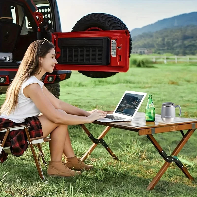 A woman sits outdoors on a wooden table working on a laptop next to an insulated stainless steel travel mug.
