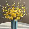 Eine Vase mit künstlichen gelben Pflaumenblütenzweigen auf einer weißen Oberfläche.