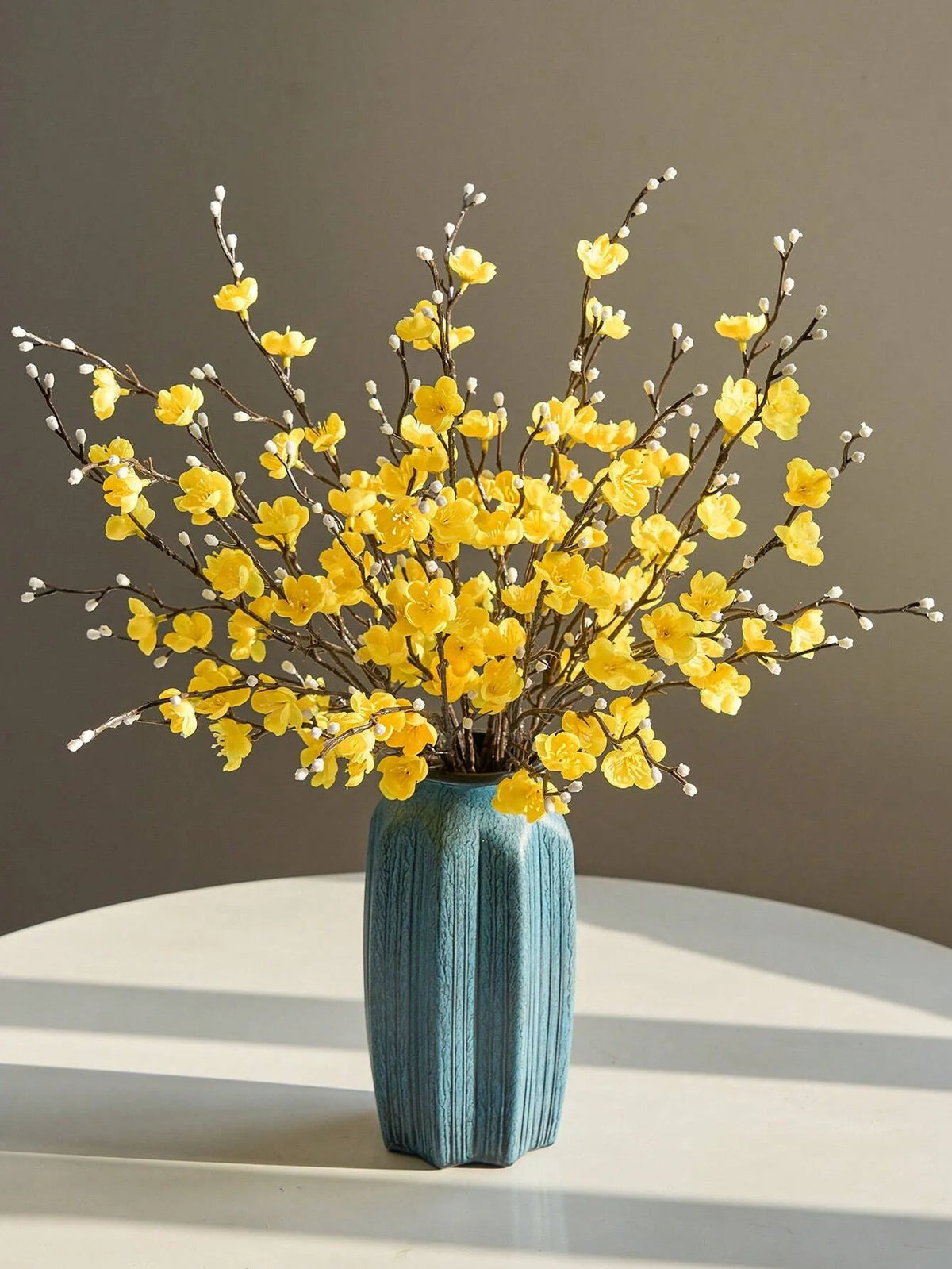 Eine Vase mit künstlichen gelben Pflaumenblütenzweigen auf einer weißen Oberfläche.
