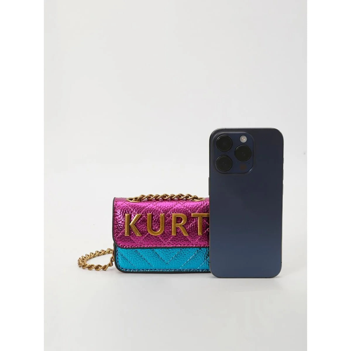 A purple and teal magnetic crossbody bag with a gold chain and letter flap next to a dark blue smartphone.