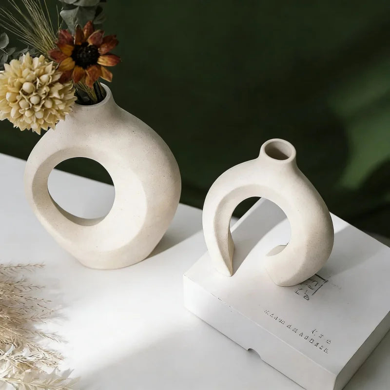 A modern minimalist ceramic vase with an organic, curved shape sits on a white surface next to dried floral elements.