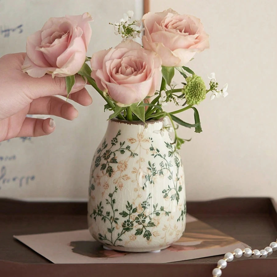 A hand holds a pink ceramic bud vase filled with light pink roses and green foliage.