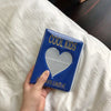 Una mano sostiene un álbum de fotos azul titulado COOL KIDS con un diseño de corazón contra una tela blanca.