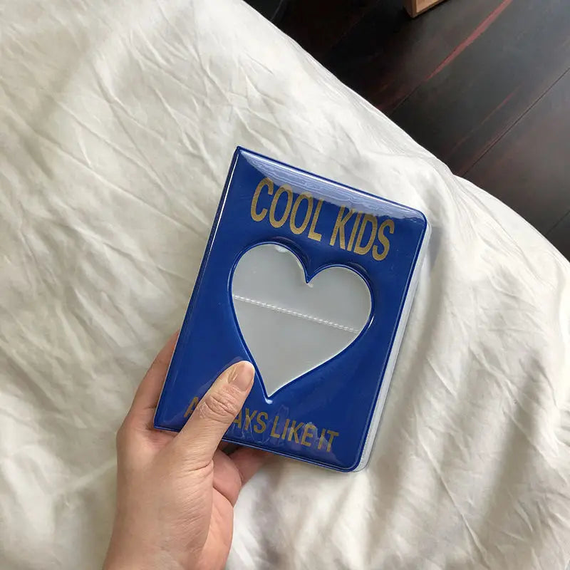 Una mano sostiene un álbum de fotos azul titulado COOL KIDS con un diseño de corazón contra una tela blanca.