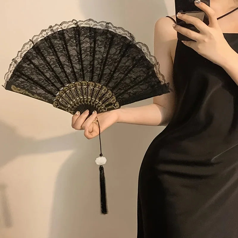 Eine Frau hält einen offenen schwarzen Spitzenhandfächer mit goldenen Akzenten vor hellem Hintergrund.