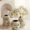 A white ceramic bud vase filled with white roses and baby's breath flowers sits on a white surface.