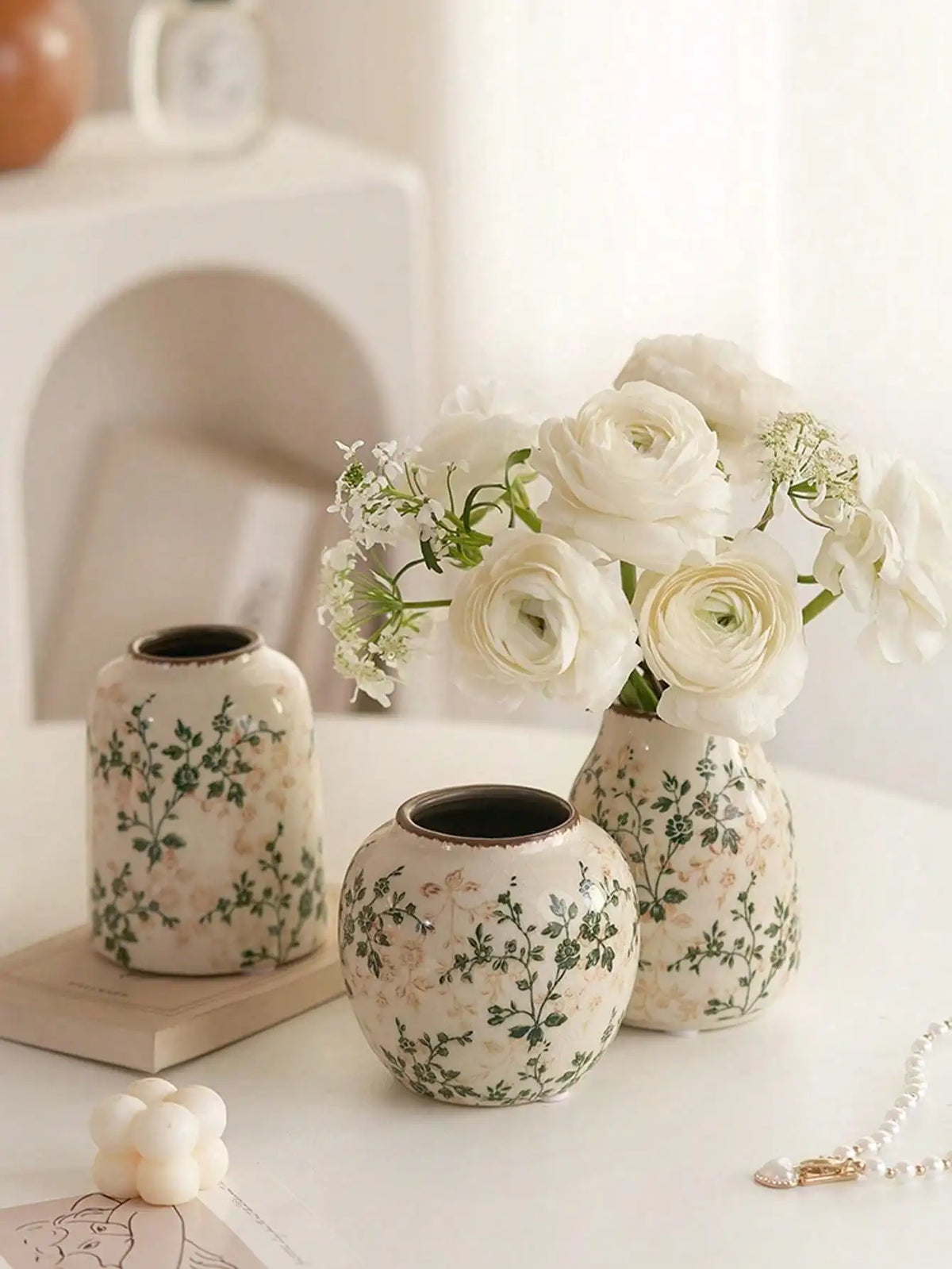 A white ceramic bud vase filled with white roses and baby's breath flowers sits on a white surface.