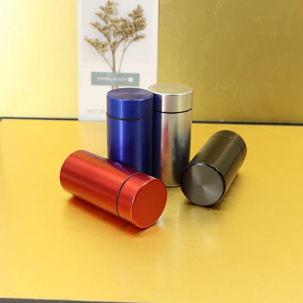 Three airtight aluminum stash jars in blue, silver, and bronze resting on a yellow surface.