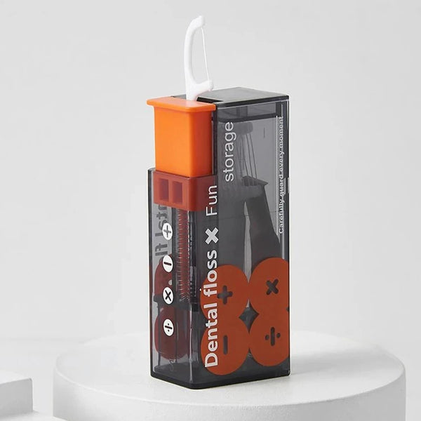 Dispenser de hilo dental negro con tapa naranja y palillos reutilizables sobre una superficie blanca.