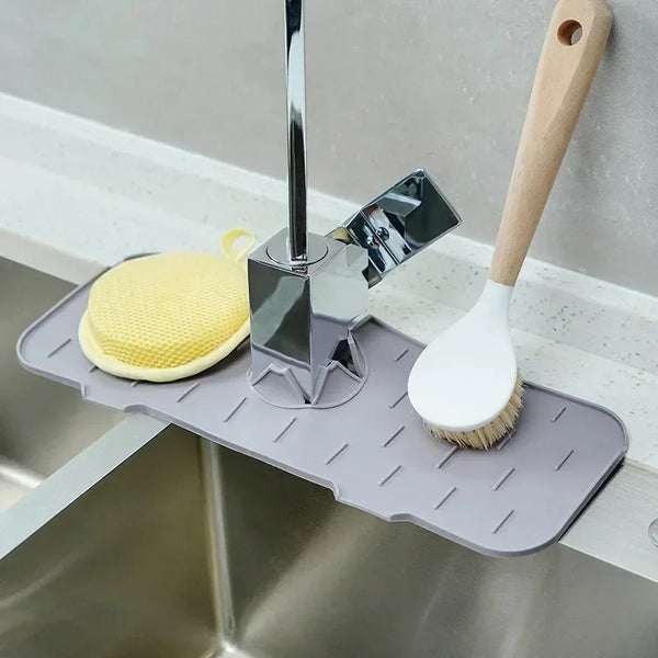 A silicone kitchen sink drainer and splash guard mat with a metal brush and wooden handle resting on a stainless steel sink.