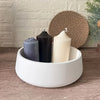 Three pillar candles in dark gray, black, and cream are displayed in a white bowl next to a woven sphere.