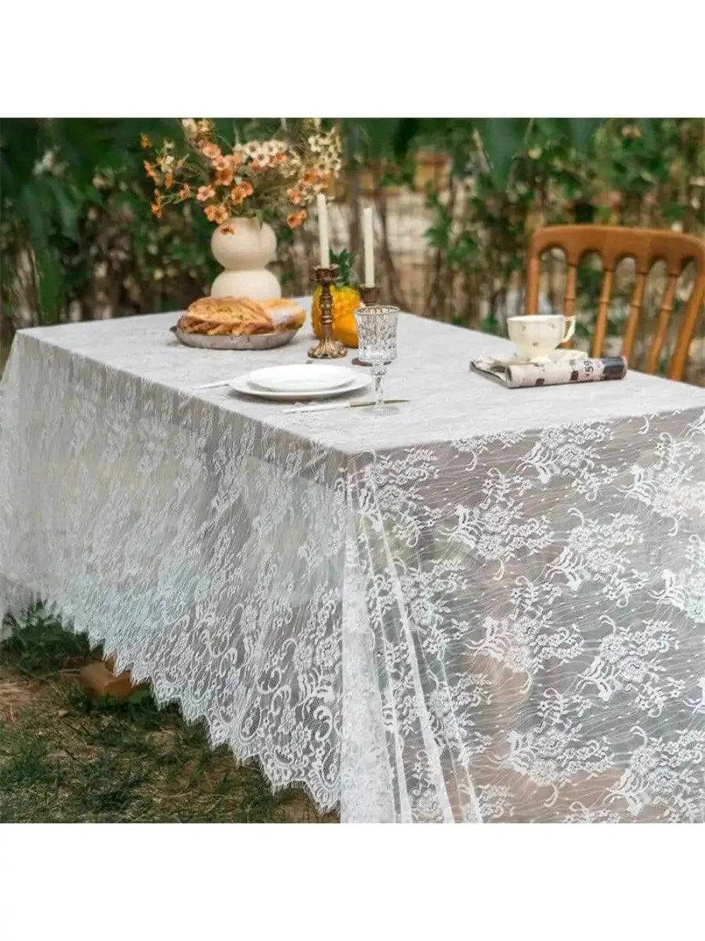 A white lace retro tablecloth is set on a table outdoors with floral decorations and food.