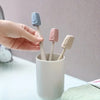 A hand holds four toothbrush head covers in various pastel colors, including blue and pink, above a white cup.