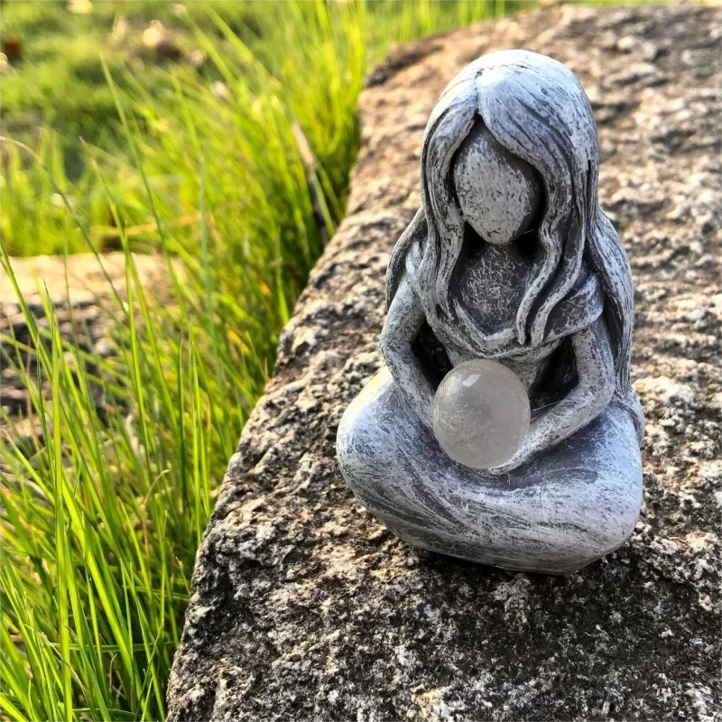 A smooth, gray resin statue of a meditating female figure with a glowing orb resting on a natural stone.