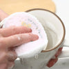 A hand is dipping a pink natural cotton cleaning sponge into a white mug.