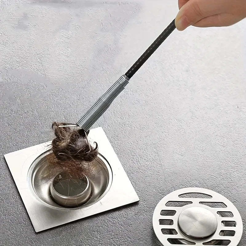 A hand is using a bendable drain auger with a claw hook to remove hair and debris from a drain.
