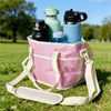 Un sac isotherme rose sur l'herbe, contenant plusieurs bouteilles d'eau réutilisables et un thermos noir.