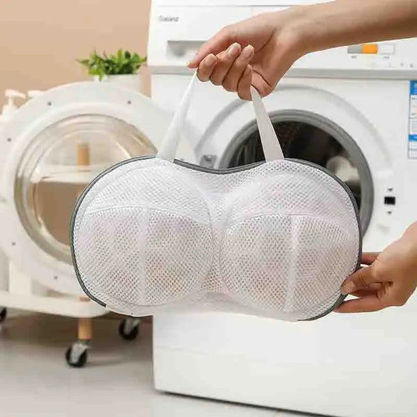 A hand is holding a white mesh laundry bag designed for bras and undergarments near a washing machine.