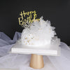 A white cake topper with gold script lettering and silver accents displayed on a white stand.