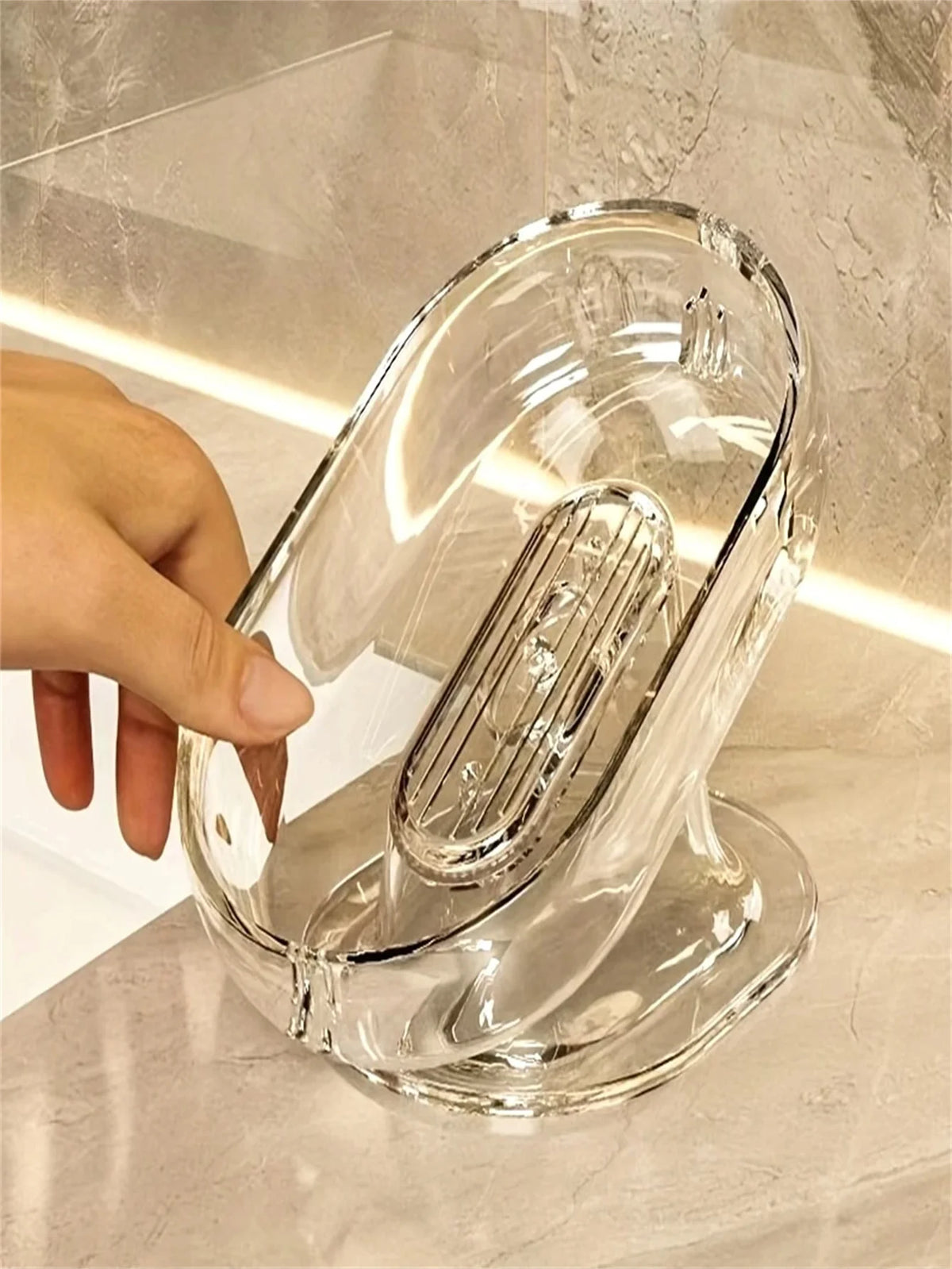 A hand is holding an elegant clear glass soap dish with a slotted design on a marble surface.