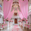 Cortinas de tela rosa drapeadas enmarcan un interior grandioso con candelabros y columnas, con pétalos rosas esparcidos en el