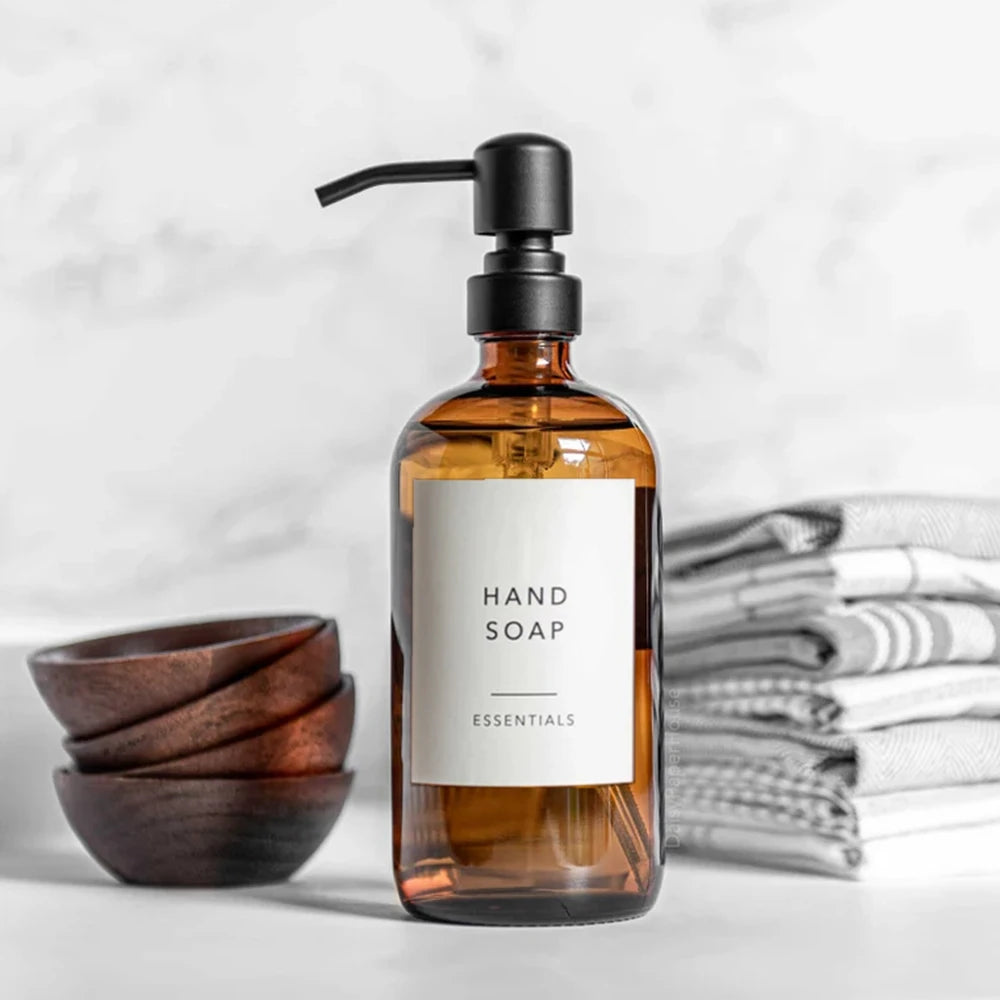 Dispenser de jabón líquido en vidrio ámbar con bomba negra, etiquetado como Hand Soap Essentials, junto a toallas blancas api